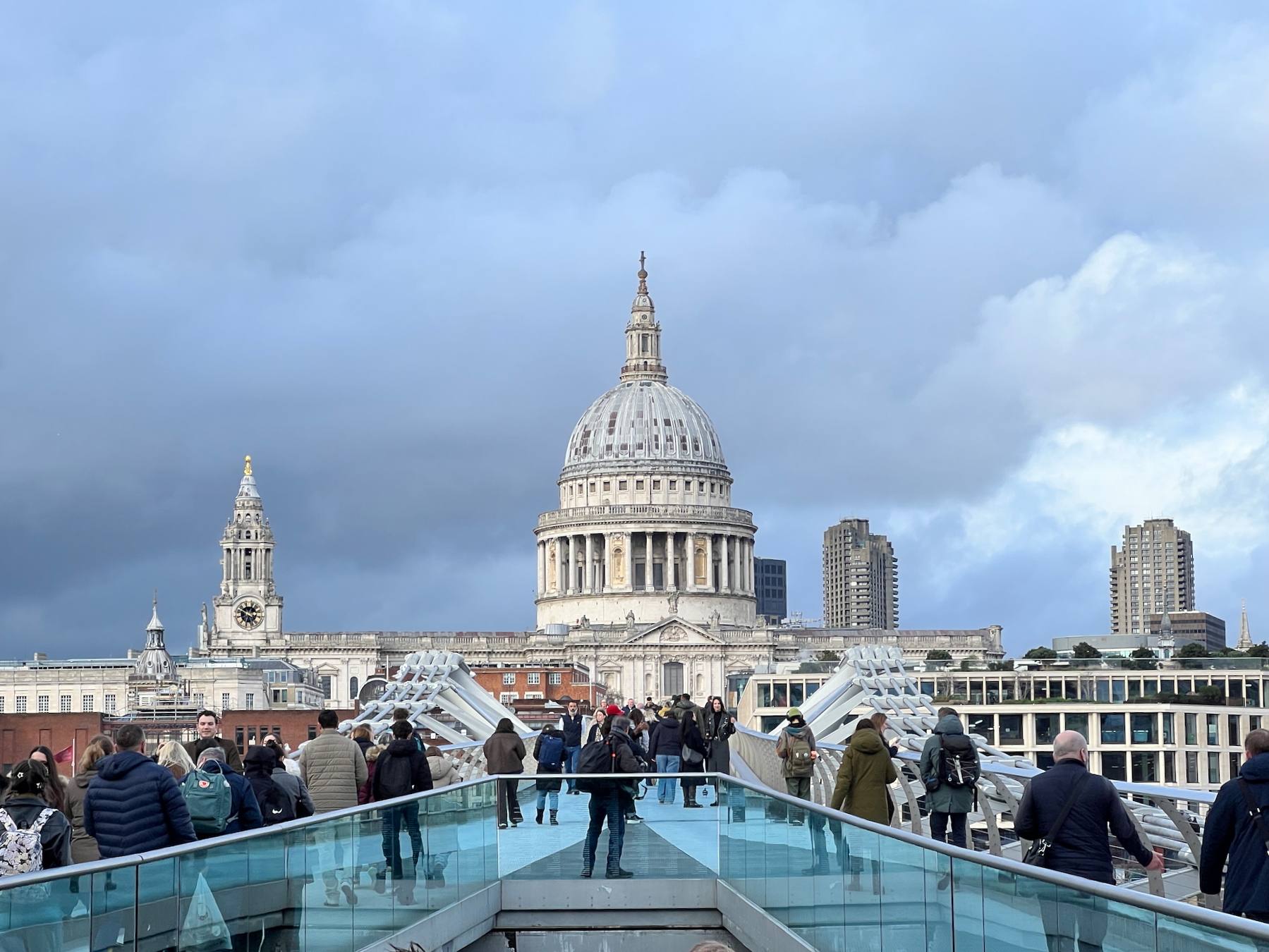Zoomed shot of large cathedral dome dominating the skyline. In the foreground people walk on a pedestrian bridge