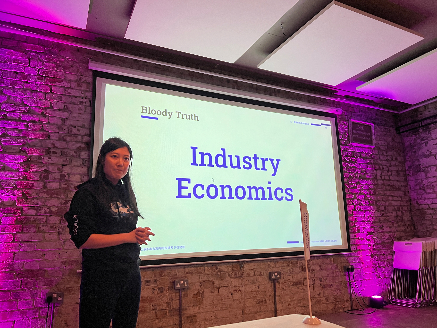 Taiwanese woman in front of a slide with the text INDUSTRY ECONOMICS and the header THE BLOODY TRUTH